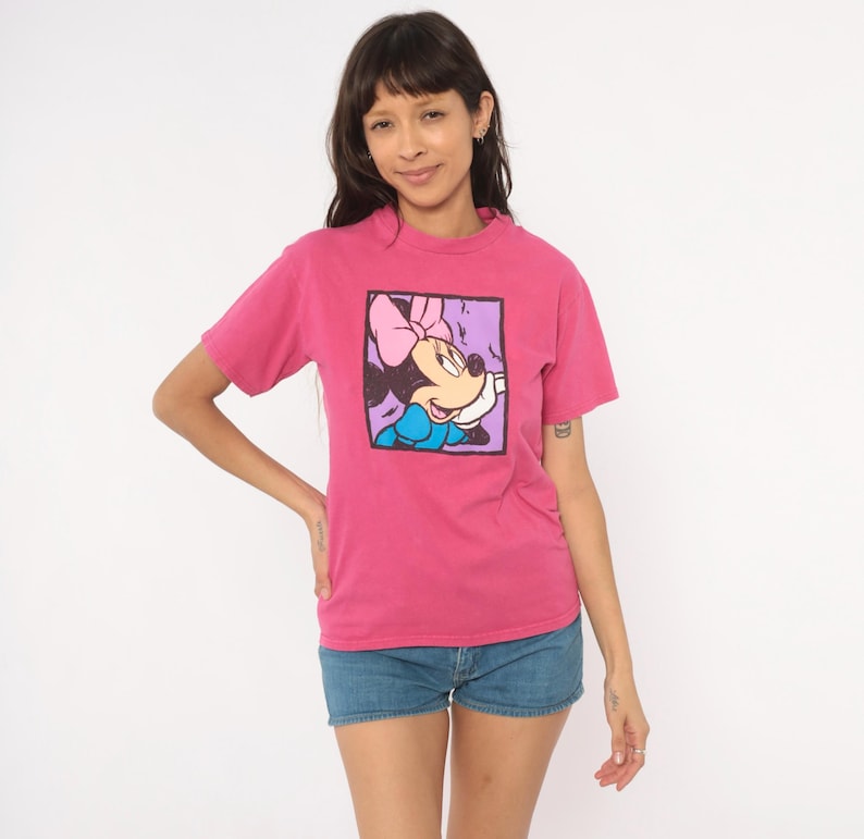 May include: A short-sleeved, crew-neck t-shirt in a vibrant magenta pink colour. The centre of the chest features a square graphic print of a cartoon mouse character wearing a pink bow and a blue dress, outlined in a thick black border.