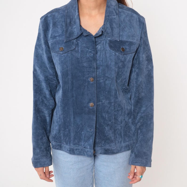 May include: A blue suede jacket with a button-up front, two chest pockets, and a collared neckline. The jacket has a slightly worn appearance and is paired with light blue jeans. The jacket's color is a medium shade of blue.