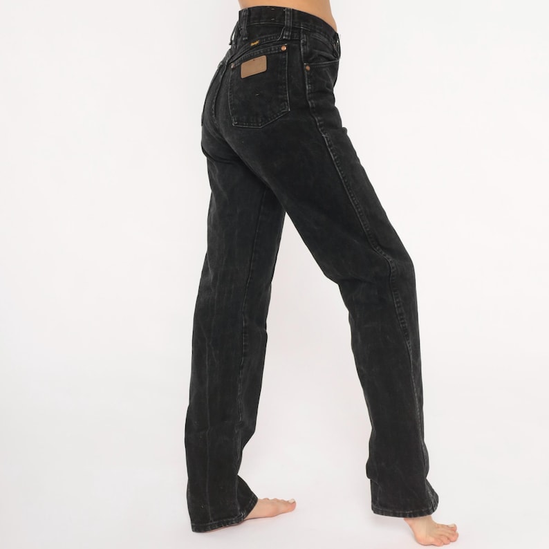 May include: Black denim jeans with a high waist and straight leg. The jeans feature a classic five-pocket design, a brown leather patch on the back, and a button and zip closure. The jeans are worn.