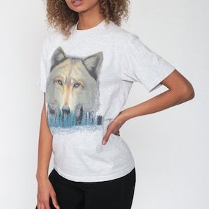 May include: A gray t-shirt with a wolf graphic. The wolf is looking directly at the viewer and is surrounded by a blue and green forest.