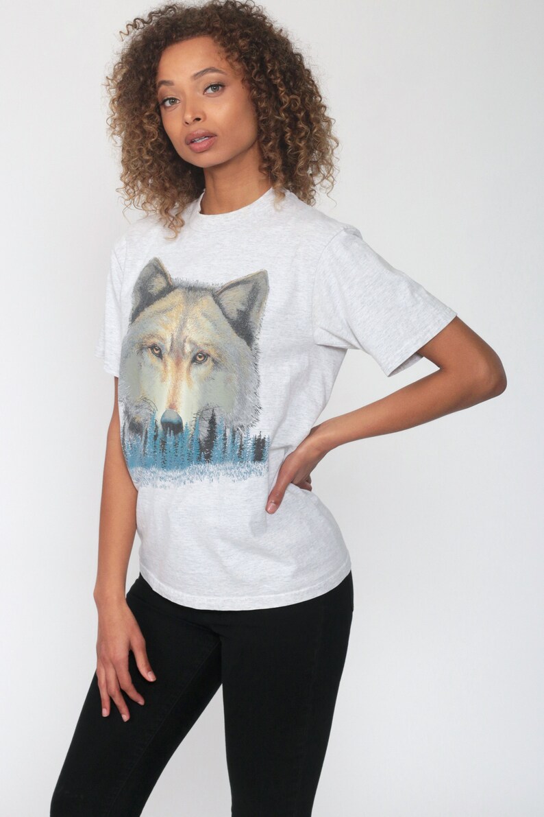 May include: A gray t-shirt with a wolf graphic. The wolf is looking directly at the viewer and is surrounded by a blue and green forest.