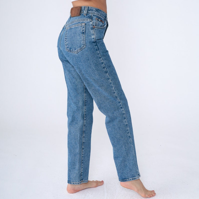 May include: Light blue denim jeans with a classic straight-leg fit. The jeans have a high waist, a button and zip closure, and a brown leather patch on the back. The jeans are a medium wash and have a five-pocket design.