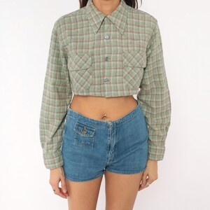 Puede incluir: Una camisa corta de manga larga con botones en un patr&oacute;n de cuadros verde salvia y rojo, con dos bolsillos en el pecho y cuello cl&aacute;sico. Combinada con pantalones cortos de mezclilla azul de talle alto con un peque&ntilde;o bolsillo frontal.