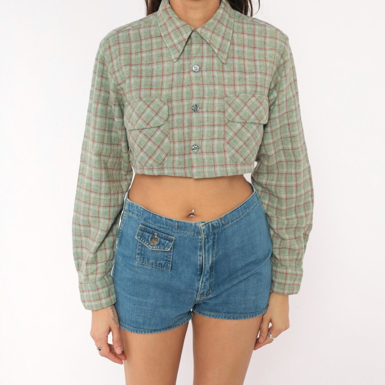 Puede incluir: Una camisa corta de manga larga con botones en un patr&oacute;n de cuadros verde salvia y rojo, con dos bolsillos en el pecho y cuello cl&aacute;sico. Combinada con pantalones cortos de mezclilla azul de talle alto con un peque&ntilde;o bolsillo frontal.