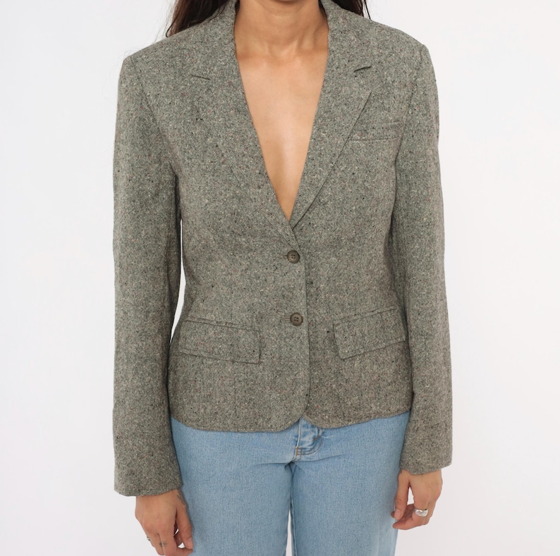 May include: A gray tweed blazer with two front pockets and two buttons. The blazer has a notched lapel and a tailored fit. The fabric has a speckled texture. The blazer is paired with blue jeans.