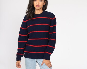 May include: Navy blue sweater with horizontal red stripes. The sweater has a crew neck and long sleeves. The model is wearing light blue jeans. The sweater is made of a soft, comfortable material.