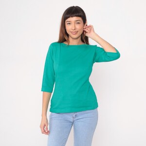 Puede incluir: Una blusa verde turquesa con mangas tres cuartos, cuello barco y corte entallado. La modelo lleva vaqueros azul claro. El fondo es blanco.