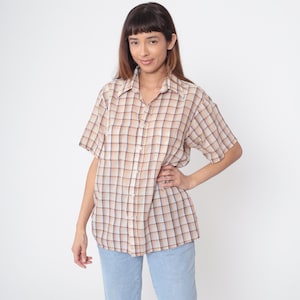 May include: A beige, brown, and blue plaid short-sleeved shirt with a collared neckline. The shirt is buttoned down and worn with light blue jeans. The style is casual and relaxed, suitable for everyday wear.