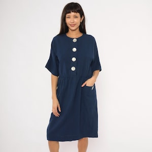 May include: A navy blue dress with large, round, silver buttons down the front. The dress has short sleeves, a cinched waist, and two front pockets. The model is wearing the dress.