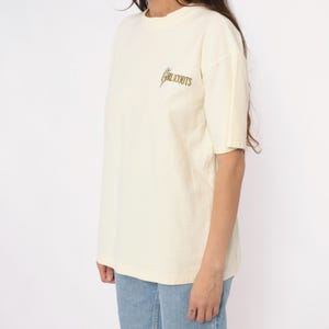 May include: Cream-colored t-shirt with short sleeves and a crew neck. The shirt features the text "GIRL SCOUTS" in a gold-colored script on the upper left chest. The model is wearing blue jeans.