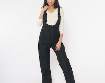 May include: Black snow bib overalls with suspenders and a front zipper. The snow pants are made of a quilted material. The person in the image is wearing a white long-sleeve shirt.