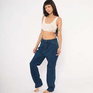 May include: A person wearing a cream-colored tank top and dark blue denim jeans. The jeans have a high waist and a relaxed fit. The person is standing in front of a white background.