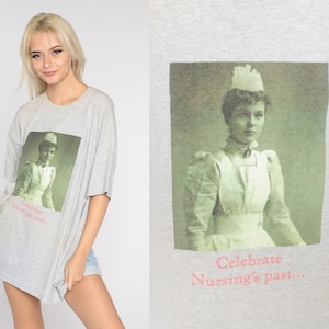 May include: A gray t-shirt with a vintage photo of a nurse wearing a white uniform and a white cap. The text on the shirt reads "Celebrate Nursing's Past..."