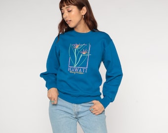 May include: A teal blue sweatshirt featuring a floral design with the word "HAWAII" in white text. The long-sleeved top is made of a soft material and is suitable for casual wear. The design includes a rectangular frame with a bird of paradise flower.