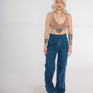 May include: A woman wearing a brown bralette with a gold chain necklace and high-waisted blue denim jeans. She has blonde hair and tattoos on her arms.
