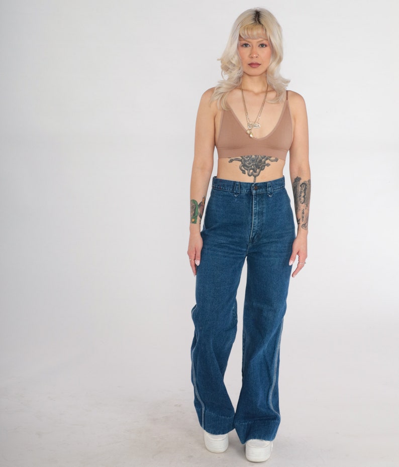 May include: A woman wearing a brown bralette with a gold chain necklace and high-waisted blue denim jeans. She has blonde hair and tattoos on her arms.