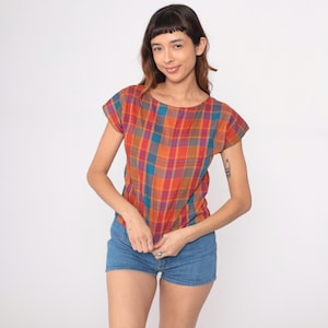 May include: A short-sleeved plaid top in shades of orange, red, teal, and purple. The top has a round neckline and a relaxed fit. The model is wearing denim shorts. The top is made of a lightweight fabric.