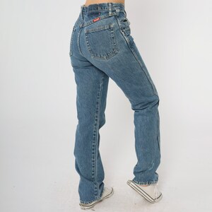 Puede incluir: Vaqueros de mezclilla azul claro con cintura alta y pernera recta. Los vaqueros tienen un parche rojo con el logo de Wrangler en el bolsillo trasero. El modelo lleva zapatillas blancas.