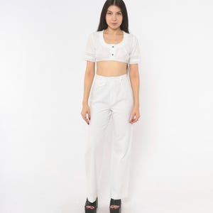 May include: A white two-piece outfit consisting of a cropped short-sleeve blouse with a scoop neckline, button-front closure, and lace trim on the sleeves, paired with high-waisted, wide-leg trousers featuring a single front pocket detail.