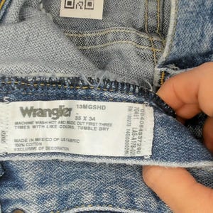 Peut inclure: Gros plan sur un jean Wrangler en denim bleu. L'&eacute;tiquette indique la marque, les informations de taille et les instructions d'entretien. Le jean est fabriqu&eacute; au Mexique avec du tissu am&eacute;ricain.