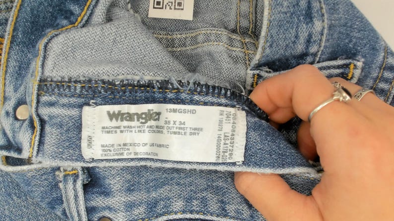 Peut inclure: Gros plan sur un jean Wrangler en denim bleu. L'&eacute;tiquette indique la marque, les informations de taille et les instructions d'entretien. Le jean est fabriqu&eacute; au Mexique avec du tissu am&eacute;ricain.