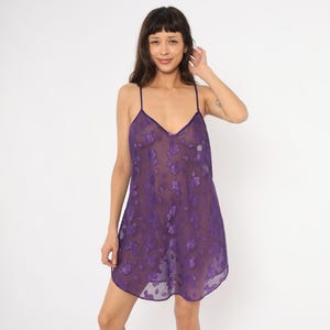 May include: A sheer purple slip dress with a V-neck and spaghetti straps. The dress is adorned with a floral pattern and has a slightly flared hemline. The fabric appears lightweight and delicate, suitable for sleepwear or lingerie.