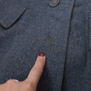 K&ouml;nnte beinhalten: Nahaufnahme eines blauen Wollmantels mit Knopfverschluss. Der Mantel hat eine Tasche und einen Kragen. Auf der Vorderseite des Mantels befinden sich braune Flecken. Ein Finger mit rotem Nagellack zeigt auf den Mantel.