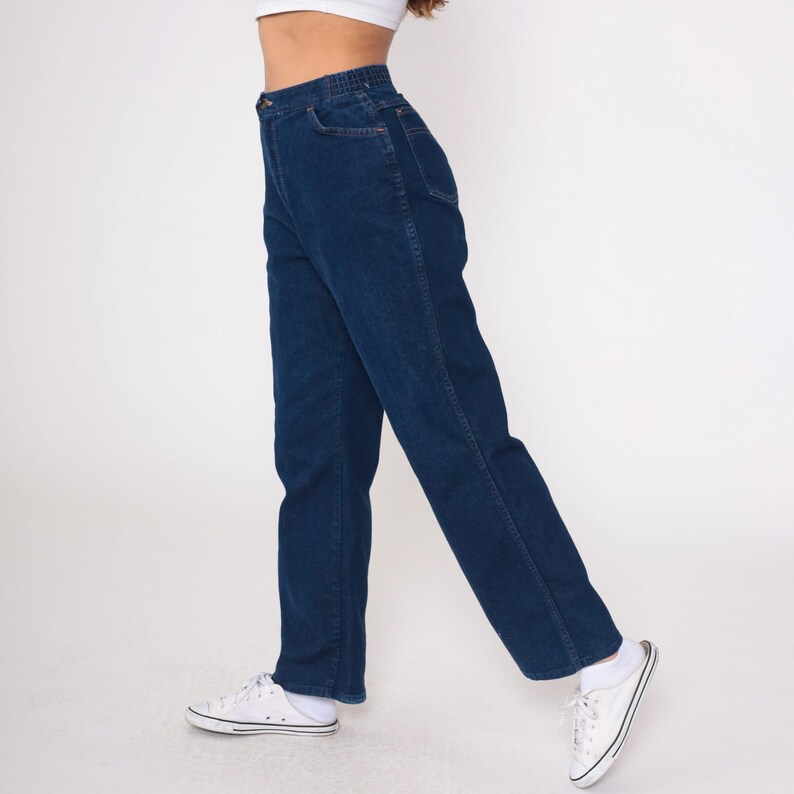 K&ouml;nnte beinhalten: Dunkelblaue Jeans mit hoher Taille und geradem Bein. Die Jeans haben ein klassisches F&uuml;nf-Taschen-Design und einen bequemen elastischen Bund. Das Model tr&auml;gt wei&szlig;e Turnschuhe.