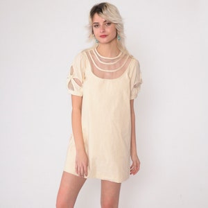 Puede incluir: Un vestido corto de manga corta de color crema con un panel de malla transparente en el escote y detalles de malla en las mangas.
