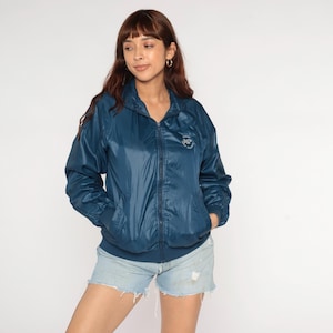 Könnte beinhalten: Türkisblaue Jacke mit Reißverschluss und kleinem Logo auf der Brust. Die Jacke hat einen Stehkragen, elastische Bündchen und eine lockere Passform. Die Jacke ist aus einem leichten, leicht glänzenden Material gefertigt. Das Model trägt Jeansshorts.