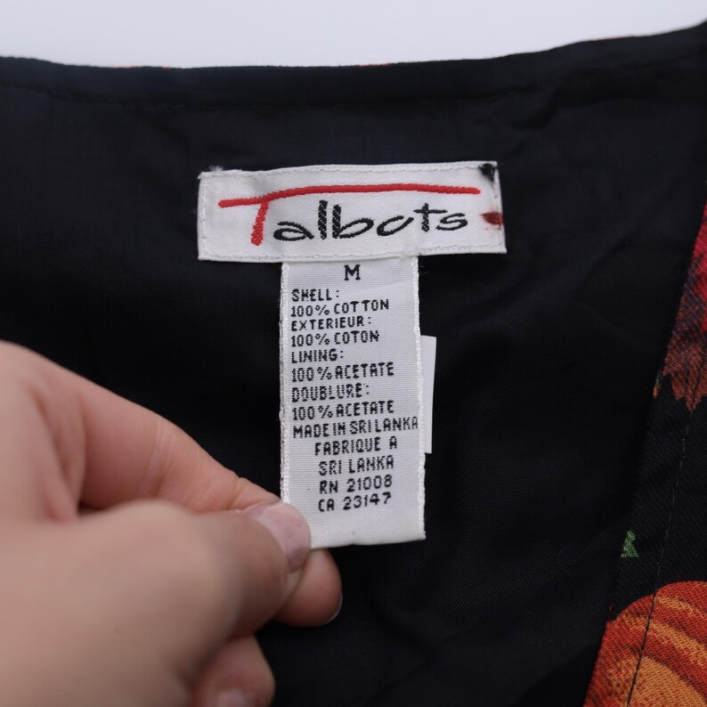 Puede incluir: Una etiqueta de ropa negra de la marca Talbots con texto blanco. La etiqueta indica que el contenido del tejido es 100% algod&oacute;n para la c&aacute;scara y el exterior, y 100% acetato para el forro y la doublure. La etiqueta tambi&eacute;n indica que la prenda se fabric&oacute; en Sri Lanka. La etiqueta incluye la talla M, RN 21008 y CA 23147.