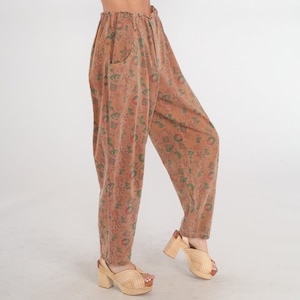 K&ouml;nnte beinhalten: Hoch taillierte Hose in ged&auml;mpftem Terrakotta-Braun mit einem Allover-Druck aus kleinen gr&uuml;nen und roten geometrischen Formen, Sonnen und abstrakten Symbolen. Die Hose hat eine lockere, zulaufende Passform mit elastischem Bund und Taschen.