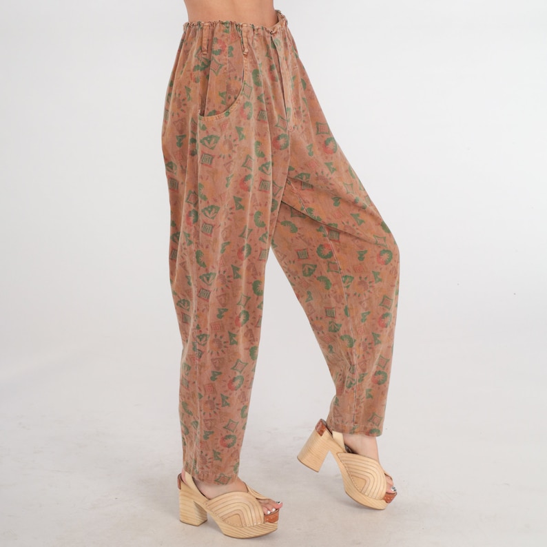 K&ouml;nnte beinhalten: Hoch taillierte Hose in ged&auml;mpftem Terrakotta-Braun mit einem Allover-Druck aus kleinen gr&uuml;nen und roten geometrischen Formen, Sonnen und abstrakten Symbolen. Die Hose hat eine lockere, zulaufende Passform mit elastischem Bund und Taschen.