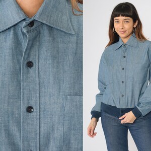 May include: A light blue denim shirt with a button-up front and long sleeves. The shirt has a collar and a pocket on the left chest.