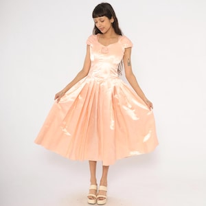 Peut inclure: Une robe en satin pêche avec un corsage ajusté et une jupe plissée ample. La robe a des manches courtes et un petit ornement floral au décolleté. Le modèle porte des sandales à plateforme.