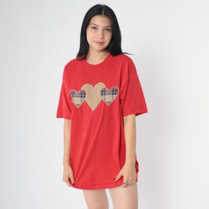 May include: A red oversized t-shirt with three plaid hearts on the front. The hearts are beige with a red, blue, and white plaid pattern.