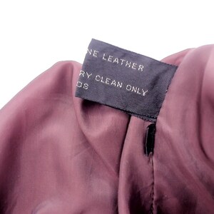 May include: Close-up of a garment's interior, revealing a dark burgundy lining. A black tag is sewn into the seam, displaying the words "GENUINE LEATHER" and "DRY CLEAN ONLY" in white text. The fabric has a smooth texture.