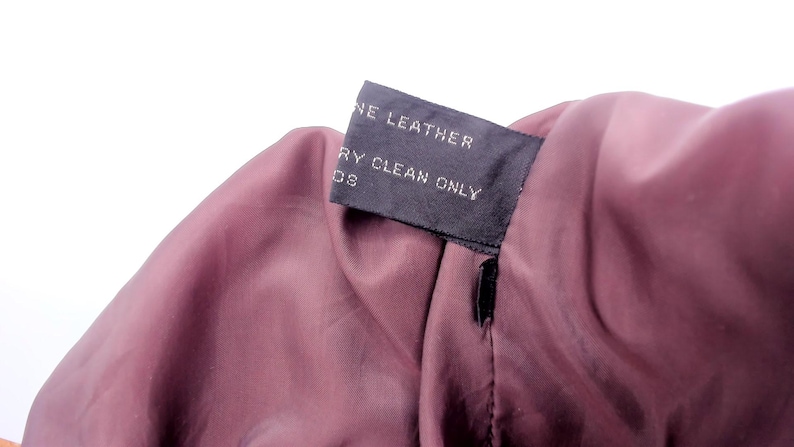 May include: Close-up of a garment's interior, revealing a dark burgundy lining. A black tag is sewn into the seam, displaying the words "GENUINE LEATHER" and "DRY CLEAN ONLY" in white text. The fabric has a smooth texture.