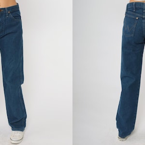 May include: Dark blue denim jeans with a classic straight leg fit. The jeans have a button and zipper closure, front and back pockets, and a brown leather patch on the back. The jeans are a medium wash and appear to be vintage.