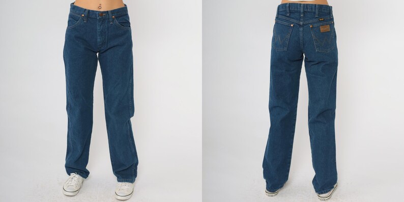 May include: Dark blue denim jeans with a classic straight leg fit. The jeans have a button and zipper closure, front and back pockets, and a brown leather patch on the back. The jeans are a medium wash and appear to be vintage.