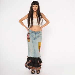May include: A light-wash denim maxi skirt with colourful patchwork details. The skirt features a black ruffled hem with floral accents. The model is wearing a white crop top and black sandals.