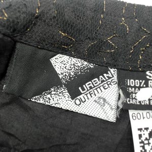 Puede incluir: Primer plano de una prenda negra con detalles dorados. La prenda tiene una etiqueta que dice "Urban Outfitters" y otra etiqueta con la talla "S" y "100% Rayon". La tela parece ser encaje.