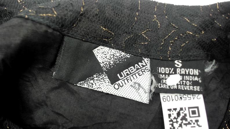 Puede incluir: Primer plano de una prenda negra con detalles dorados. La prenda tiene una etiqueta que dice "Urban Outfitters" y otra etiqueta con la talla "S" y "100% Rayon". La tela parece ser encaje.