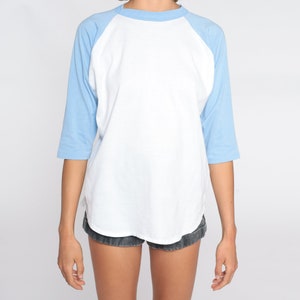 May include: A white and light blue baseball style t-shirt. The shirt features a white body with light blue sleeves and a light blue crew neck. The sleeves are three-quarter length. The shirt is made of a soft, comfortable material.