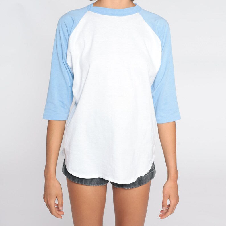 May include: A white and light blue baseball style t-shirt. The shirt features a white body with light blue sleeves and a light blue crew neck. The sleeves are three-quarter length. The shirt is made of a soft, comfortable material.