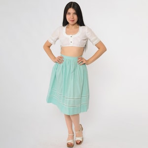 Könnte beinhalten: Ein weißes Crop-Top mit Spitzenbesatz und drei Knöpfen, kombiniert mit einem türkisfarbenen Vichy-Rock. Der Rock ist knielang und hat einen passenden Spitzenbesatz. Das Outfit ist im Vintage-Stil.