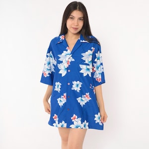 May include: A short-sleeved button-down shirt in a vibrant blue color featuring a repeating floral pattern. The flowers are rendered in shades of white and light blue with subtle pink accents. The shirt has a classic collar and a single chest pocket.