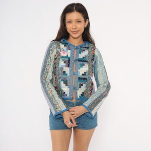 May include: A long-sleeved quilted jacket featuring a patchwork design with various floral and geometric patterns in shades of blue, white, and pink. The jacket has a round neckline, blue trim, and a front closure with four small fabric-covered buttons.