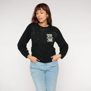 May include: Black long-sleeved jumper featuring a sparkly teddy bear motif on the chest. The jumper has a round neckline and is decorated with small, sparkly embellishments across the top. The sleeves are long and the fit is relaxed.