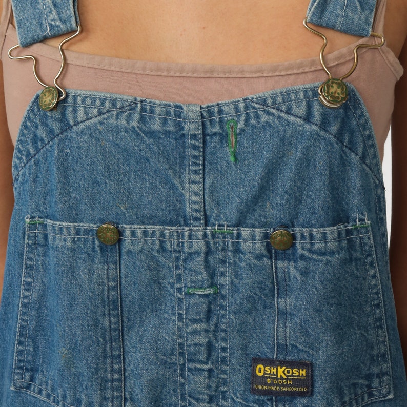 May include: Blue denim dungarees with green stitching detail and a brown OshKosh B'Gosh logo patch. The dungarees have adjustable straps with metal buckles.